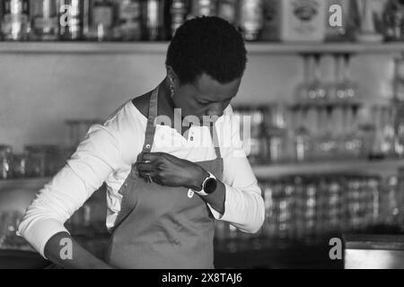 Il barista concentrato che indossa un grembiule si trova di fronte a una varietà di bottiglie in un bar pieno Foto Stock