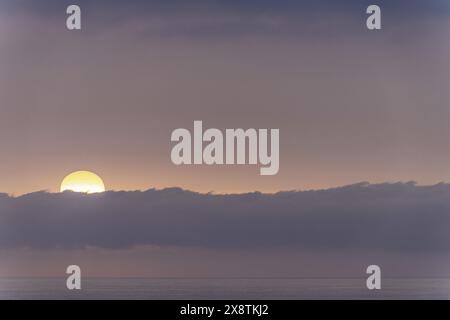 Una serena alba sull'orizzonte dell'oceano, con il sole parzialmente nascosto da una bassa nuvola. I delicati colori pastello del cielo creano un ambiente tranquillo e rilassante Foto Stock