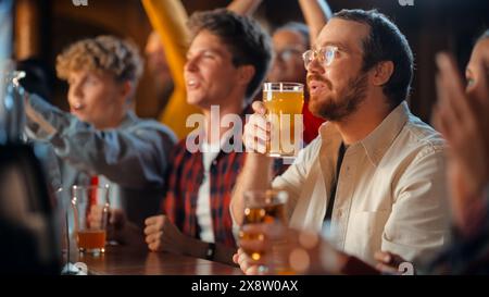 Ritratto di un giovane felice che celebra una vittoria allo Sports Pub durante una partita di calcio. Guardando la TV mentre sorseggi birra a un banco, diventa emotivo dopo che la squadra segna un gol. Foto Stock