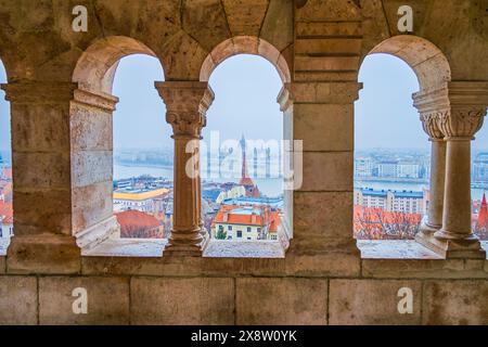 La vista attraverso le finestre della galleria del Bastione dei pescatori, che mostra il Parlamento, le case e il fiume Danubio a Budapest, Ungheria. Foto Stock