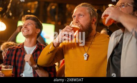 Gruppo di buoni amici che si divertono in un pub sportivo. Tre uomini che fanno il tifo per la loro squadra di calcio preferita. I giovani celebrano e brindano i bicchieri di birra quando i giocatori segnano un gol e vincono. Foto Stock