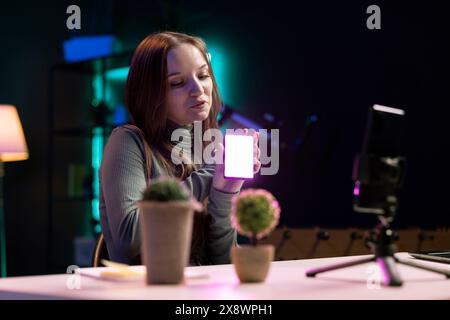 Gen Z girl in studio di appartamenti che utilizza il telefono cellulare per registrare, rivedendo luci mini RGB. Il giovane intrattenitore filma l'elettronica con il cellulare sul treppiede, presentando un dispositivo di illuminazione a LED Foto Stock