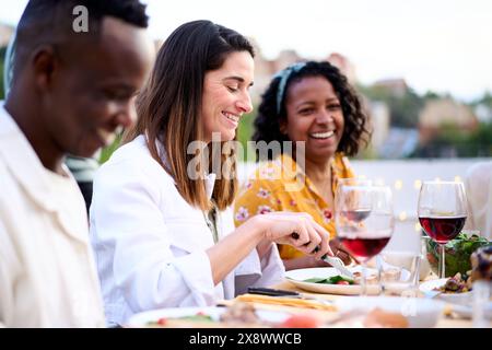 Vista laterale di una donna caucasica sorridente che mangia cibo seduto a tavola con gli amici non concentrati sul tetto. Foto Stock