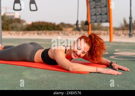 Donna rossa sorridente sdraiata su un tappetino da yoga. Una donna allegra in abbigliamento sportivo che fa esercizio nel parco estivo all'aperto. Concetto di fitness e salute. Foto Stock