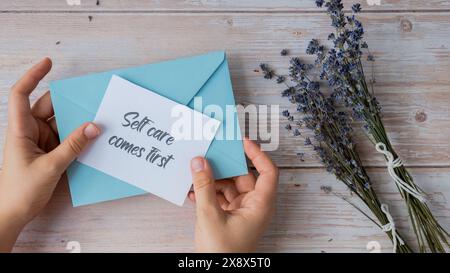 L'AUTOASSISTENZA VIENE PRIMA DI TUTTO il testo su un promemoria cartaceo di supporto dalla busta blu. Composizione piatta fiori asciutti di lavanda. Concetto di felicità interiore, rallentare la disintossicazione digitale e la realizzazione personale. Vista dall'alto Foto Stock