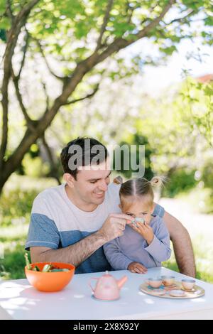 Papà tratta la sua piccola figlia con un tè da una tazza giocattolo mentre si siede a un tavolo in giardino a fare una festa Foto Stock