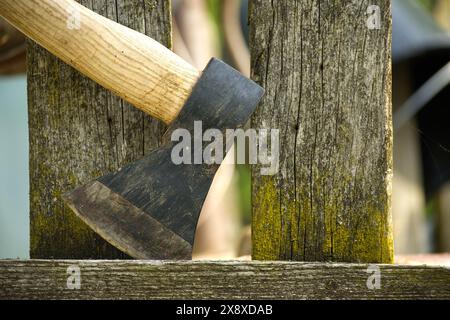 Vista ravvicinata di un'ascia, caratterizzata da un manico marrone e da una lama grigio scuro, alloggiata saldamente in un vecchio palo di recinzione in legno resistente agli agenti atmosferici con incrinature visibili Foto Stock