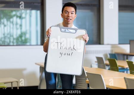 Un giovane studente universitario maschio sta tenendo un cartello con scritto "grazie" su una lavagna. Sta sorridendo alla telecamera e sta in piedi in un cla Foto Stock