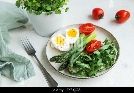 Una sana insalata primaverile si trova su un tavolo bianco, con uova bollite a fette, asparagi verdi vivaci e pomodori ciliegini maturi insieme a rucola fresca Foto Stock