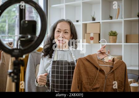 Un'attraente venditrice online asiatica matura sta mostrando i suoi prodotti in diretta sui suoi social media, parlando con i suoi clienti in streaming e vendendo cl Foto Stock