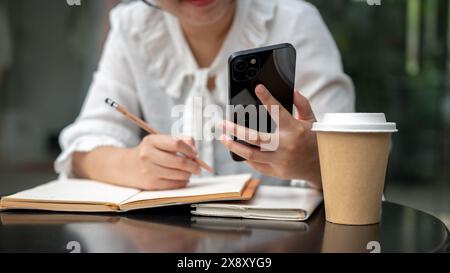 Un'immagine ritagliata di una giovane donna asiatica positiva che usa il suo smartphone mentre scrive qualcosa nel suo notebook a un tavolo. fare i compiti, fare i compiti Foto Stock