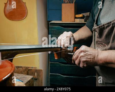 Primo piano delle mani di un abile artigiano che ripara delicatamente le corde di un violino in un laboratorio di liuteria rustico e ben attrezzato Foto Stock