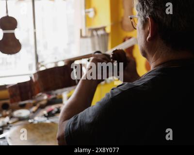 Vista a spalla di un liutaio che perfeziona una chitarra, con attenzione allo strumento e alle mani in un ambiente accogliente e soleggiato Foto Stock