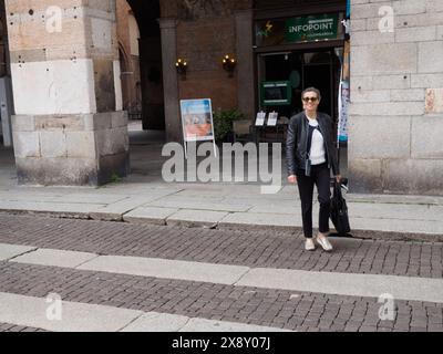 Cremona, Italia - 15 maggio 2024 donna moderna con occhiali da sole e giacca in pelle si trova in una piazza urbana vicino all'ingresso di un infopoint Foto Stock