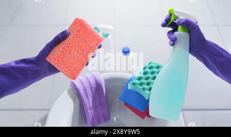 Mani femminili che indossano guanti in gomma e tengono in mano prodotti per la pulizia, WC sullo sfondo, shot POV Foto Stock