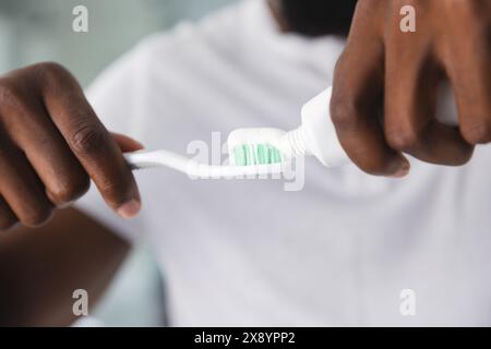 A casa, un afroamericano che applica il dentifricio sullo spazzolino da denti. Indossare una camicia bianca, concentrandosi sul mantenimento di una buona igiene dentale, inalterata Foto Stock