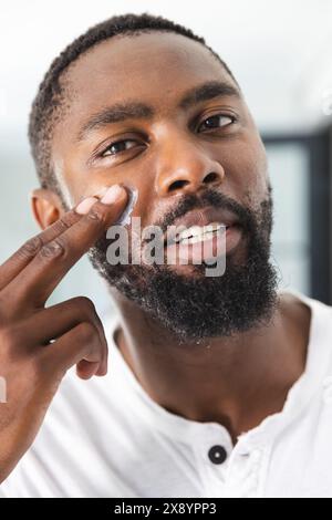 A casa, un uomo afroamericano che applica la crema sotto gli occhi Foto Stock