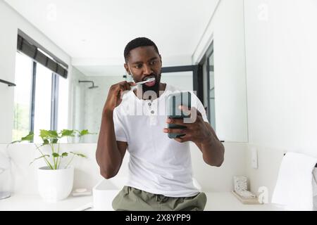 A casa, un uomo afroamericano che pulisce i denti, controlla il telefono Foto Stock