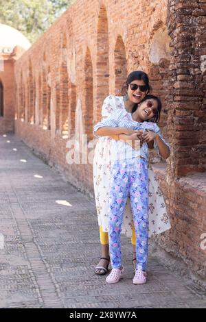 Madre e figlia indiane che indossano occhiali da sole si divertono insieme vicino a un antico muro di mattoni Foto Stock