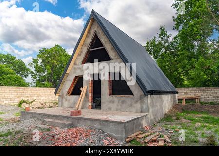 Una piccola casa con un muro di mattoni e un tetto e' in costruzione. Il tetto ha una forma triangolare. Foto Stock