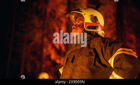 Coraggioso giovane combattente afroamericano dei vigili del fuoco con l'uniforme di sicurezza e un casco che guarda intorno alla foresta durante un incendio. Leader di squadra professionale che valuta la pericolosa situazione ecologica. Foto Stock