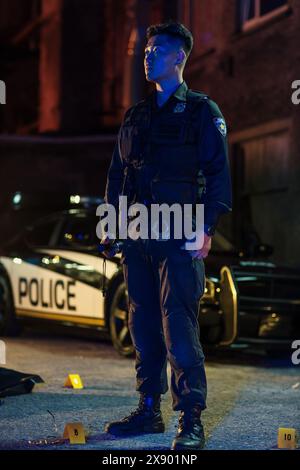 Ritratto dell'agente di polizia asiatica in una scena del crimine di notte. Colpo a corpo intero di un giovane poliziotto in uniforme di mantenimento della legge e dell'ordine in città sotto la sirena della polizia di Neon Foto Stock