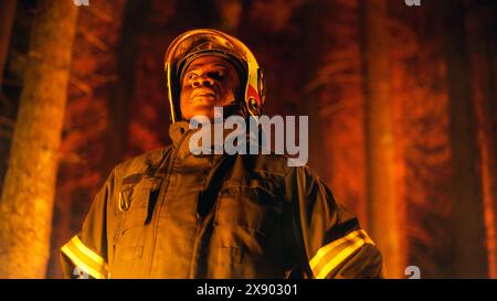 Coraggioso giovane combattente afroamericano dei vigili del fuoco con l'uniforme di sicurezza e un casco che guarda intorno alla foresta durante un incendio. Leader di squadra professionista che valuta la situazione pericolosa. Foto Stock