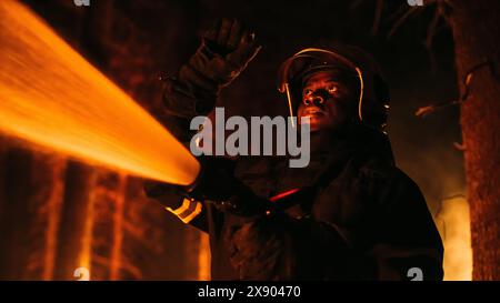 Primo piano Ritratto creativo Low Angle di un coraggioso combattente dei vigili del fuoco afroamericano che usa una manichetta per combattere un incendio boschivo. Black Fireman tiene in mano il tubo dell'acqua ad alta pressione, combattendo contro il fuoco boschivo. Foto Stock