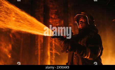 Primo piano Ritratto creativo Low Angle di un coraggioso combattente dei vigili del fuoco afroamericano che usa una manichetta per combattere un incendio boschivo. Black Fireman tiene in mano il tubo dell'acqua ad alta pressione, combattendo contro il fuoco dei boscaioli. Foto Stock