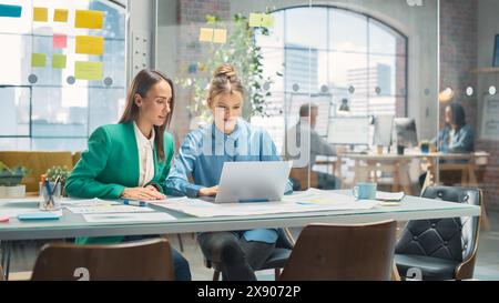 Due colleghi creativi che discutono e indicano un notebook in un ufficio casual moderno. Responsabile vendite caucasico che discute il piano di progetto con l'assistente legale femminile e prende appunti Foto Stock
