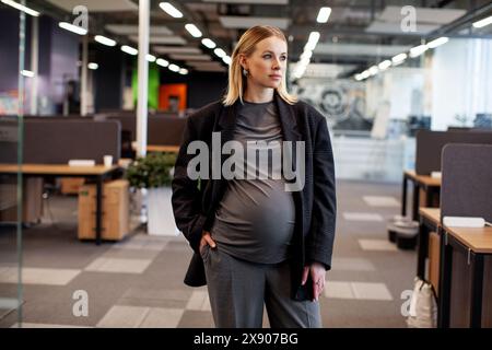 Bella donna d'affari incinta in ufficio, vestita con un elegante abbigliamento da lavoro per la maternità composto da un blazer e pantaloni, che mettono in mostra professionisti Foto Stock