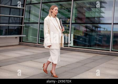 Bella donna bionda incinta con un abito da lavoro bianco con un blazer e un vestito, che cammina all'aperto con un laptop vicino a un edificio aziendale Foto Stock