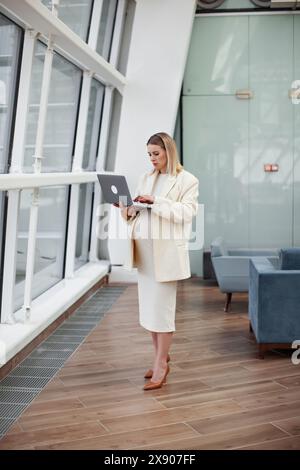 Donna d'affari incinta che lavora con sicurezza su un notebook in un moderno ufficio con sfondo azzurro e indossa un abito elegante e un blazer, re Foto Stock