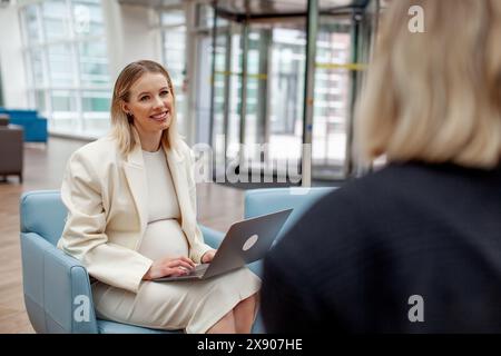 Donna incinta in tuta bianca durante una riunione d'affari con la collega in un moderno ufficio o in un centro di coworking. Donna d'affari professionista che lavora in multitasking Foto Stock