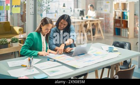 Ritratto di due giovani colleghi creativi che si consultano l'un l'altro utilizzando un notebook nell'ufficio moderno. Supervisore ispanico maschile che parla di pianificazione con il capo operativo bianco femminile. Foto Stock