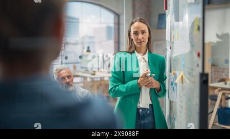Una donna Project Manager realizza una presentazione per un team creativo diversificato nella sala riunioni di un'agenzia. I colleghi si riuniscono attorno al tavolo della conferenza e discutono di sviluppo e progettazione aziendale. Foto Stock