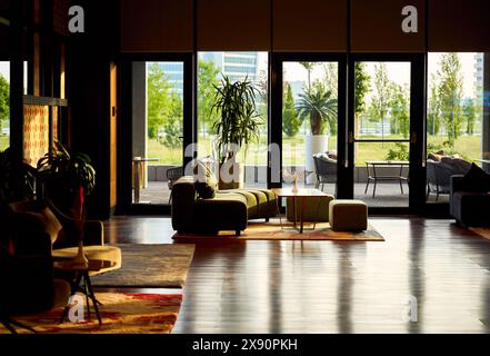 Moderna lobby di lusso con finestra panoramica, piccolo tavolo moderno in legno, piantatrice con cespugli verdi e accogliente divano con riflessi sul pavimento. Foto Stock