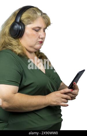 Questa fotografia cattura una donna bianca caucasica di mezza età immersa nel mondo digitale attraverso il suo smartphone mentre indossa le cuffie Bluetooth. Foto Stock