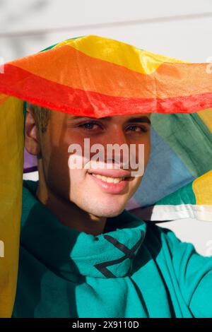 Un giovane latino che indossa un maglione verde con una bandiera arcobaleno sorride. La bandiera è sulla sua testa, è all'aperto al tramonto. Foto Stock