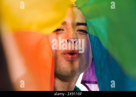 Vista frontale ravvicinata di Latin Gay Man che guarda la telecamera che lancia un bacio coperto da una bandiera d'orgoglio. Concetto di umorismo e leggerezza, come l'uomo è ma Foto Stock