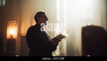 Nella Chiesa cristiana: Il Ministro guida la Congregazione nella preghiera, legge dal Santo Libro, dalla Bibbia, dal Vangelo di Gesù. Il ritratto del sacerdote dà speranza alle persone fedeli. Ripresa cinematografica drammatica Foto Stock