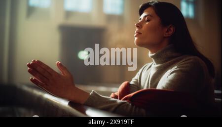 La giovane donna cristiana siede con pietà nella Chiesa maestosa, con le mani piegate cerca la guida della fede e della spiritualità mentre prega. Fede religiosa nel potere e nell'amore di Dio Foto Stock