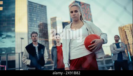 Ritratto di una bella giovane donna sportiva che guarda la fotocamera e tiene in mano una palla da calcio rossa. Elegante calciatrice donna in piedi dietro una recinzione con un gruppo di persone multiculturali Foto Stock