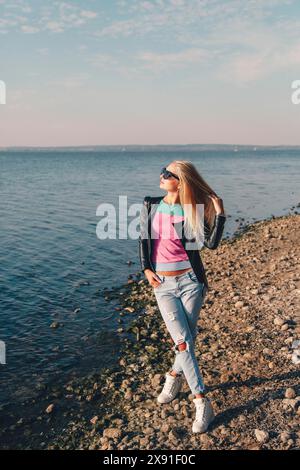 Donna con occhiali da sole e outfit casual in piedi su una spiaggia rocciosa vicino all'acqua in una giornata di sole, Bielorussia, Minsk Foto Stock