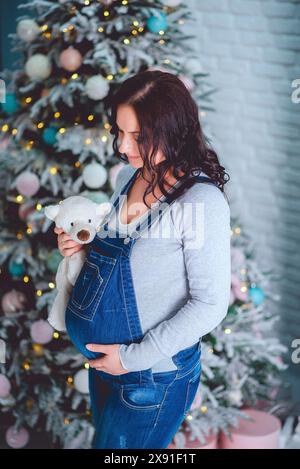 Donna incinta in tuta di denim che tiene in mano un peluche di orso vicino a un albero di Natale decorato con luci e baule colorate Foto Stock