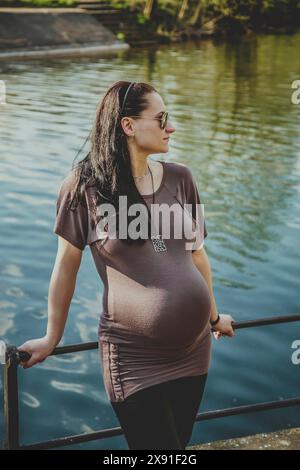 Una donna incinta che indossa occhiali da sole e un abbigliamento casual si trova accanto a una ringhiera vicino all'acqua, riflettendo un'atmosfera tranquilla. Bielorussia, Minsk Foto Stock