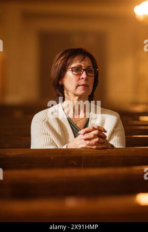 Una devota donna cristiana anziana siede piamente in una Chiesa, cerca una guida dalla sua fede religiosa e spiritualità. Spirito del cristianesimo e fede nella bontà di Dio, stringendo le mani per pregare, Foto Stock