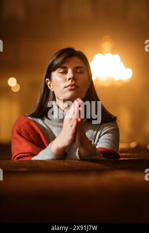 La donna cristiana siede piamente nella Chiesa, pregando, cerca la guida e il conforto dalla fede religiosa e dalla fede spirituale in Dio. Si concentra sulla sua spiritualità con gli occhi chiusi e le mani di preghiera Foto Stock