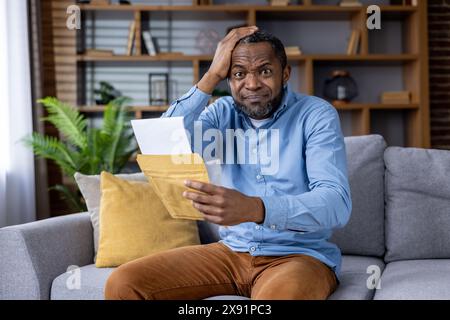 In un soggiorno, un uomo siede su un divano, sembra scioccato mentre legge una lettera sorprendente. Il suo linguaggio del corpo riflette la sua reazione emotiva alle notizie inaspettate che ha appena ricevuto a casa Foto Stock