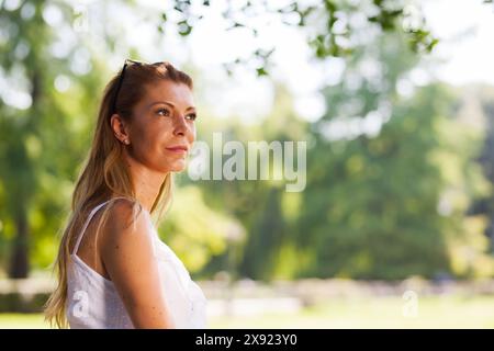 Una donna premurosa di mezza età che indossa un abito bianco contempla tranquillamente in un lussureggiante parco verde inondato dalla luce del sole. Foto Stock